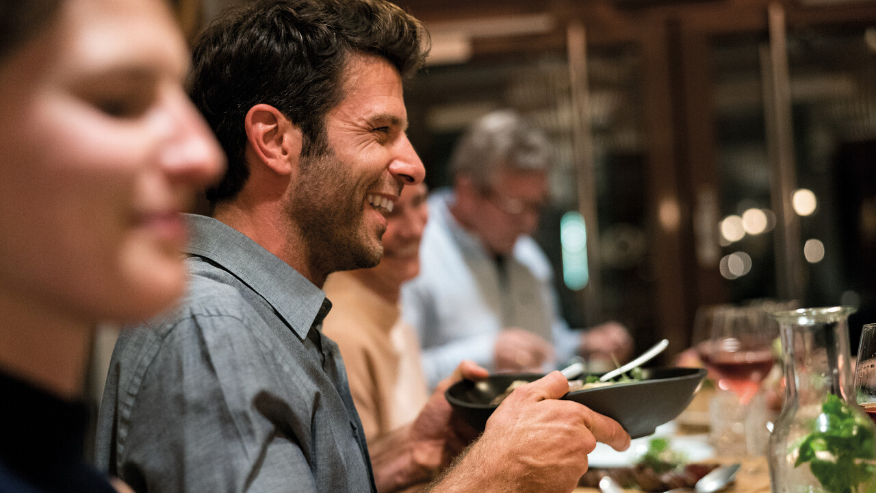 StrandGut Resorts glueckliche Gaeste essen im StrandGut Resort in St. Peter-Ording zu Abend, junger Mann haelt Salatschale