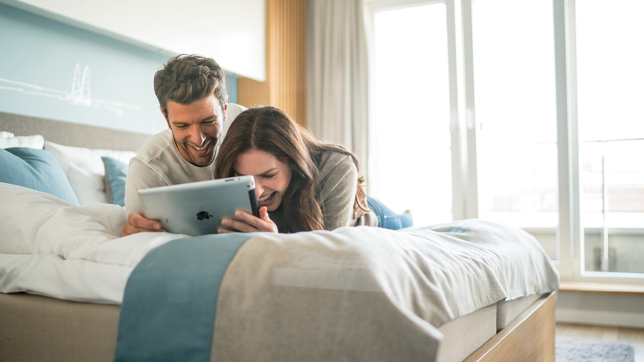 StrandGut Resorts junges Paar liegt lachend auf einem Bett und schaut auf ein Ipad