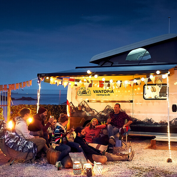 Vantopia Camping Vans Keyvisual Vans am Strand mit Lichteketten und jungen Urlaubern die gemeinsam die Nacht genießen