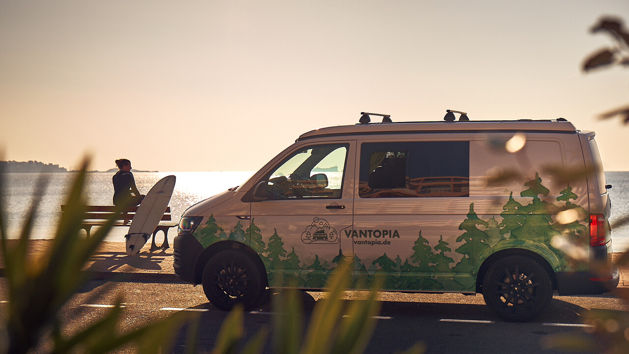Vantopia Dreamer Van mit Frau im Neoprenanzug und Surfbrett auf einer Bank am Strand